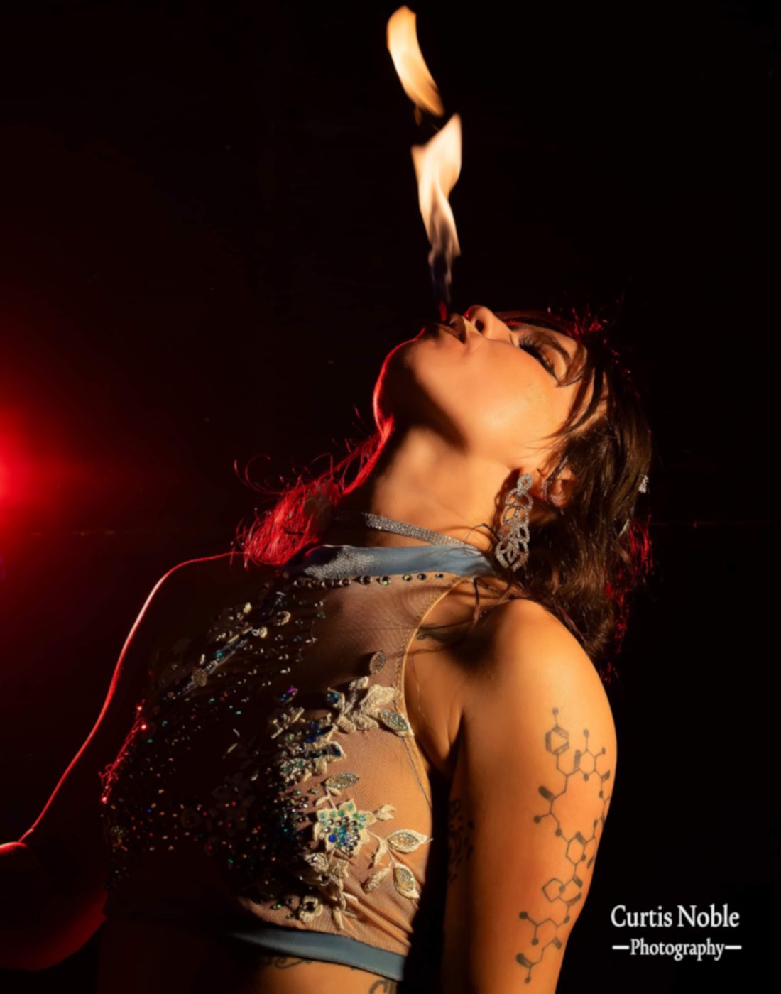 Darby demonstrates fire breathing during a fire dancing performance wearing an ornate costume with her head tilted back while blowing a flame out of her mouth.